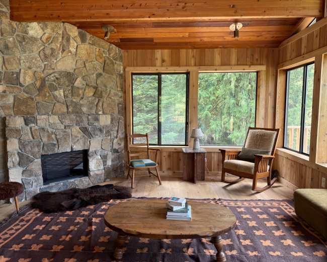 The interior of a wooden cabin features a stone fireplace, large windows offering views of greenery, and rustic furniture arranged around a central wooden coffee table.