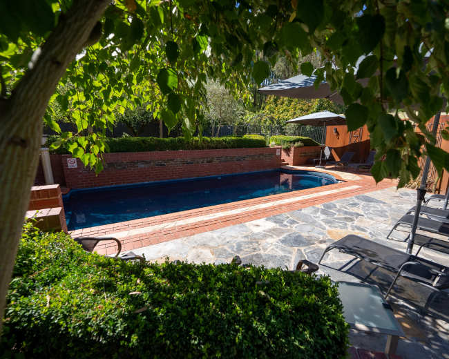 The image shows a backyard scene featuring a rectangular pool surrounded by stone pavers, lounge chairs, and lush greenery.