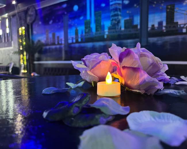 A small candle sits surrounded by delicate flower petals on a dark table, with a mural of a city skyline in the background.