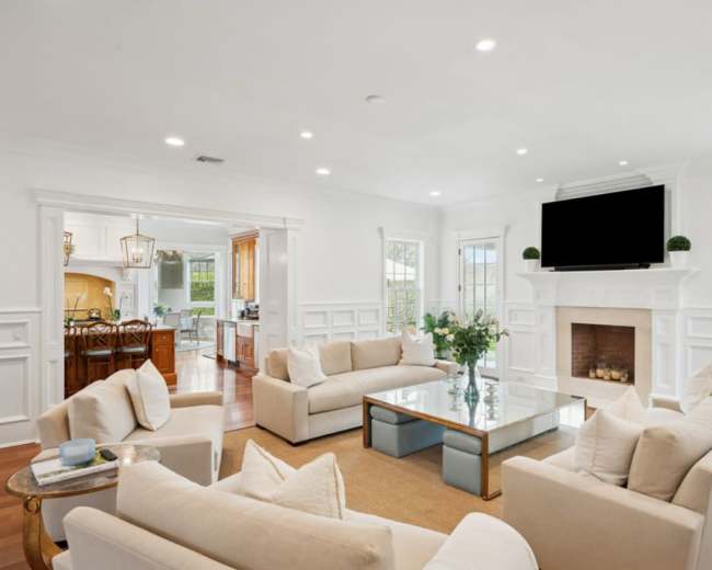 The image features a bright, modern living room with two beige sofas, a glass coffee table, a fireplace, and large windows leading to an outdoor area.