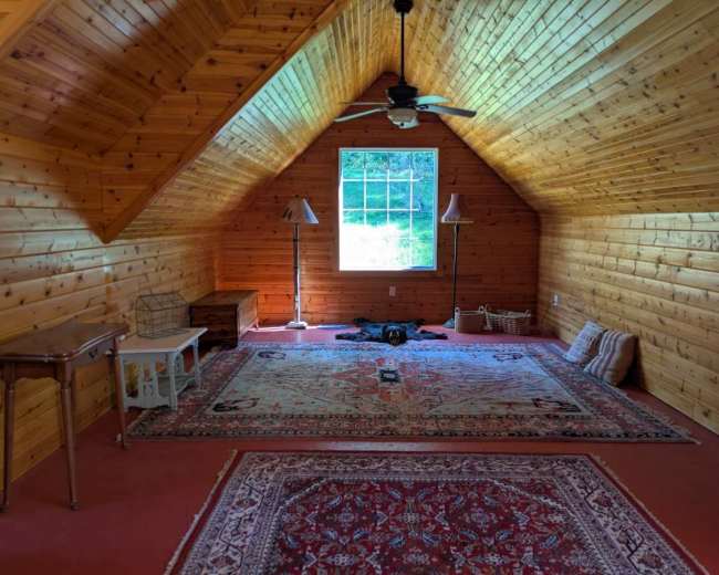 The image shows a spacious attic room with wooden walls and a sloped ceiling, featuring a window, a rug, a table, and a relaxed dog on the floor.