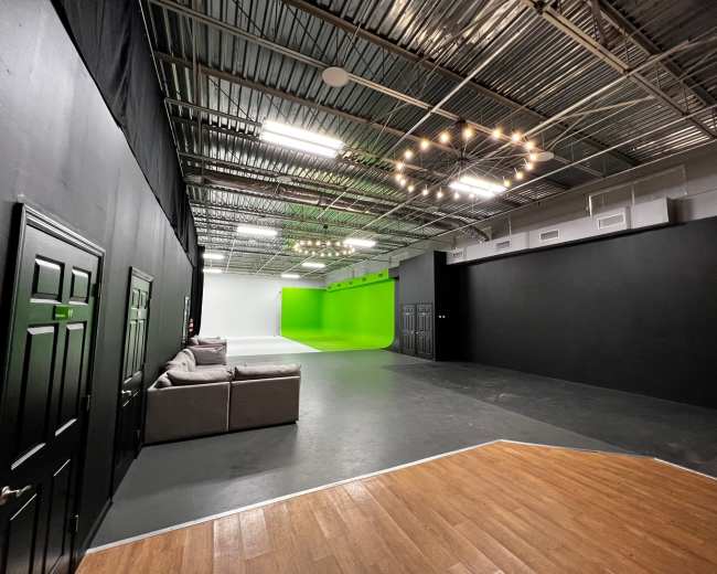 The image shows a spacious studio with gray flooring and black walls, featuring a green screen area at one end and a seating area with a couch.