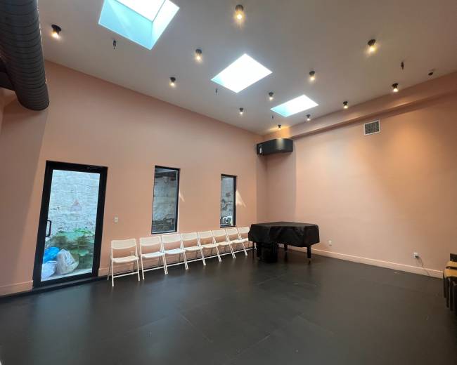 The image shows a spacious room with pink walls, a black table in the center, and several white chairs lined up against one wall, illuminated by ceiling lights and skylights.