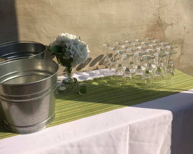 The image shows a table setup featuring two metal buckets, a floral arrangement, and a row of clear glassware on a green-striped tablecloth against a textured wall.
