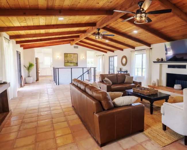 The image shows a spacious living room with a high, wooden ceiling, featuring a seating area with leather sofas, a coffee table, and a fireplace.