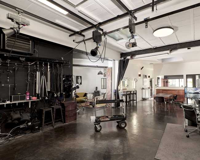 The image shows a modern photography studio equipped with lighting fixtures, a cart on wheels, and multiple workstations with computers.