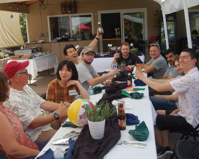 A group of ten people sits around a long table covered with colorful decorations under a large canopy in a backyard setting.