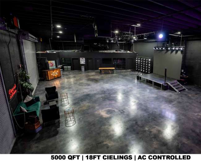 The image shows a spacious, empty event venue with a polished concrete floor, a small stage with lighting, and seating areas against the walls.