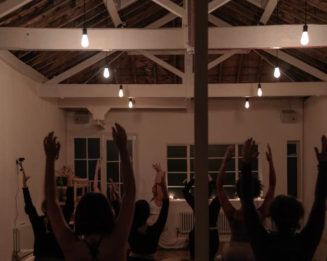 A group of people is practicing yoga in a dimly lit studio with wood beams and low-hanging lights.