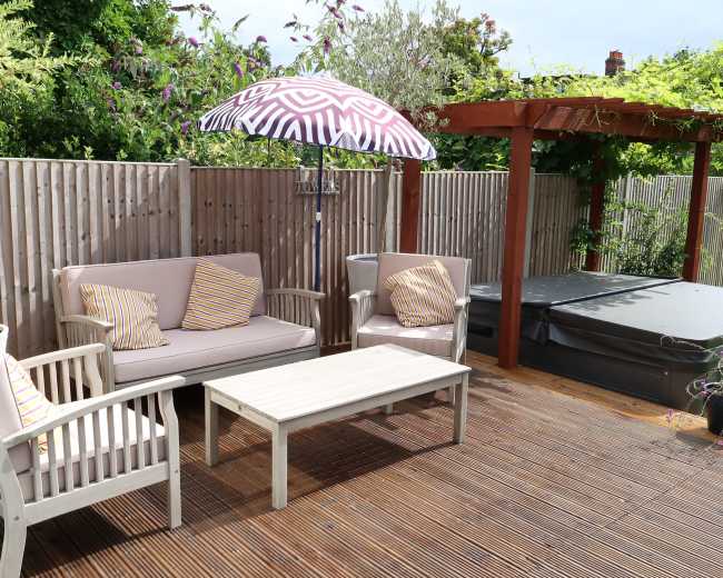 A patio area features a seating arrangement with cushions, a table, an umbrella, and a hot tub surrounded by a wooden fence and greenery.