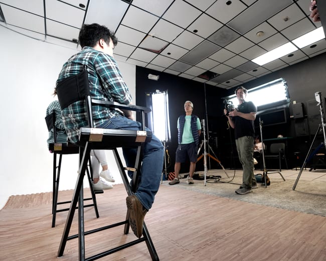 A small film studio setup features two individuals seated on high chairs while a crew member operates a camera, and others prepare lighting equipment.