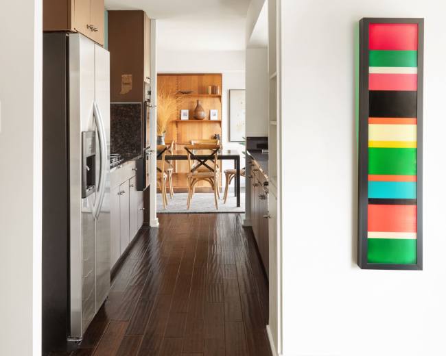 A modern kitchen features stainless steel appliances and a colorful wall art piece, leading to a dining area with a wooden table and chairs.