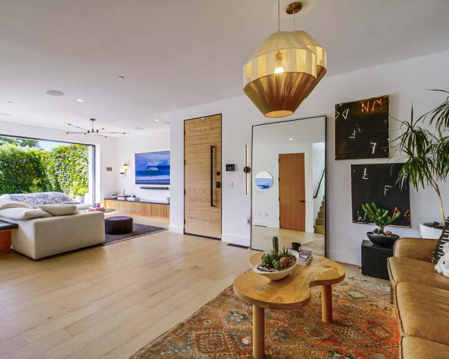 The image shows a modern living room with large windows, wooden flooring, and a blend of contemporary furniture and decor.