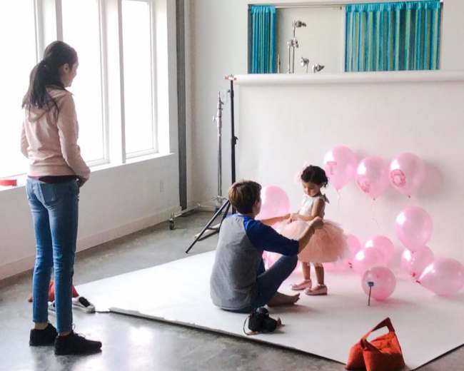 A child in a pink dress is being adjusted by an adult in a photo studio with pink balloons arranged in the background.