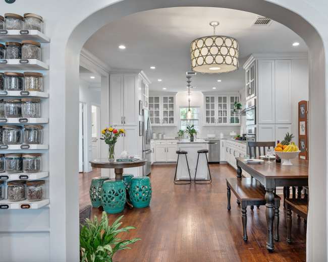 A spacious kitchen features a large island, a wooden dining table, and walls lined with jars, framed by an arched entrance.