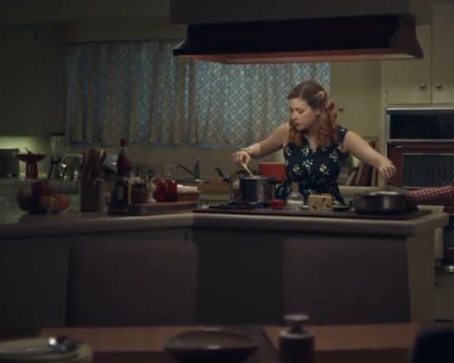 A woman cooks at a kitchen island while several pots are arranged on the stove, with a colorful fridge and scattered kitchen items in the background.