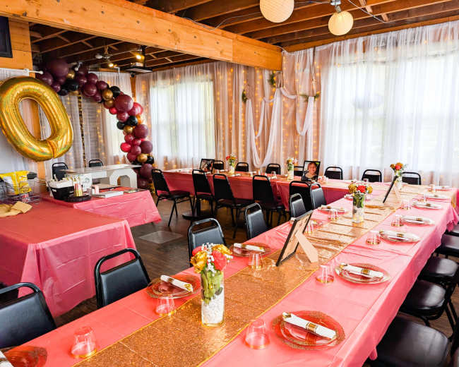 The image shows a decorated event space with tables covered in pink tablecloths, a long glittery centerpiece, balloons in various colors, and floral arrangements.