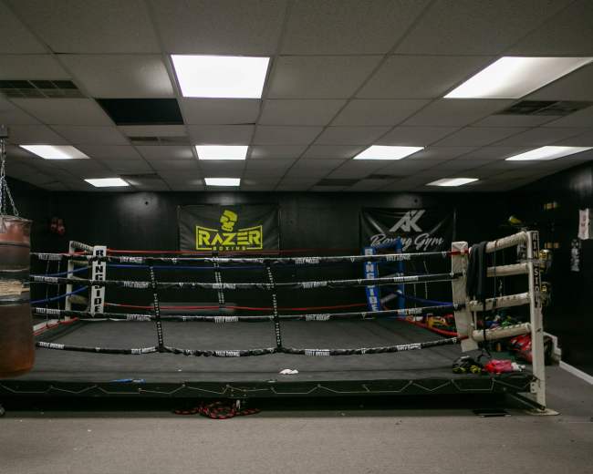 The image shows a boxing gym with a ring in the center, surrounded by various boxing equipment and advertisements on the walls.