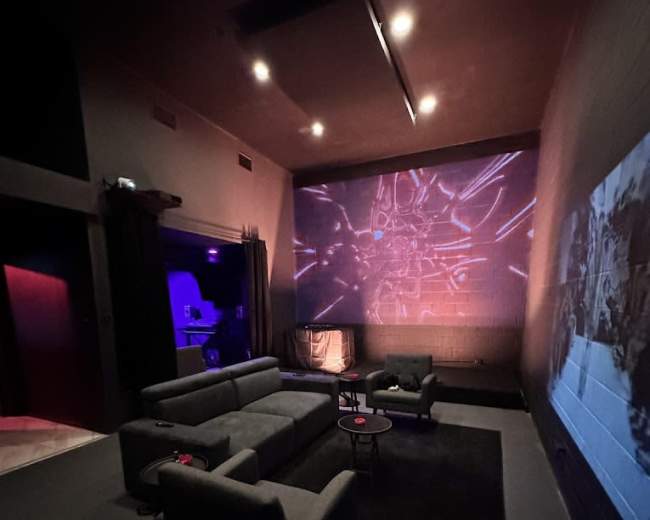 The image shows a dimly lit lounge with gray sofas, a low table, and a wall displaying a bright projection.