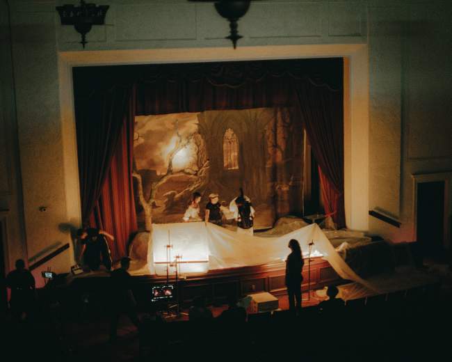 A group of people prepares a stage with a painted backdrop, draped in white fabric, in a theater setting.