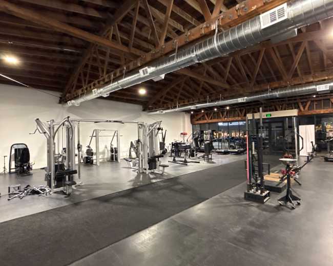 The image shows a modern gym interior with various exercise equipment, including weight machines, free weights, and workout stations, under a wooden beam ceiling.