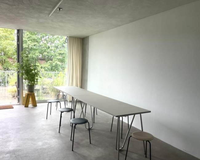 The image shows a minimalist room with a long table and several chairs, facing a large window that overlooks greenery.