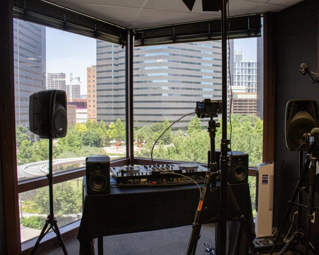 The image shows a modern recording studio setup with a DJ mixer and speakers positioned by a large window overlooking a cityscape and greenery.