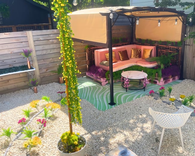 The image shows a landscaped outdoor space featuring a covered seating area with pink cushions, a round table, and decorative plants arranged on gravel.