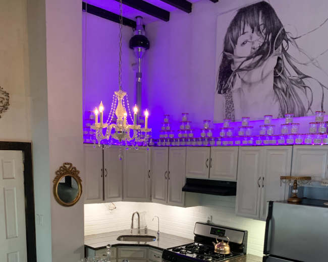 The image shows a kitchen with gray cabinets, a silver refrigerator, and a black and white checkered floor, featuring a chandelier, a decorative tablecloth, and a large wall mural.