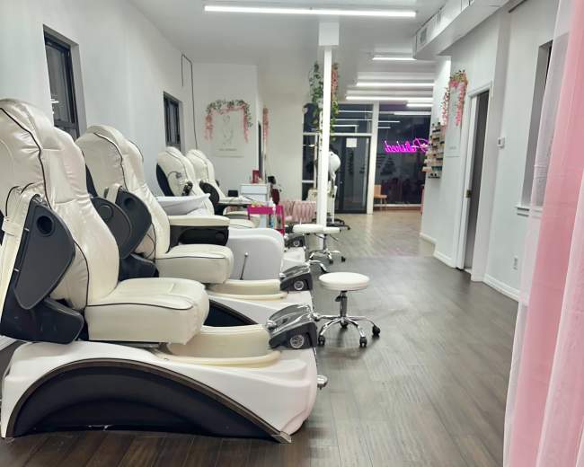 The image shows a nail salon interior with several ergonomic pedicure chairs arranged along one side and a rolling stool in the foreground.