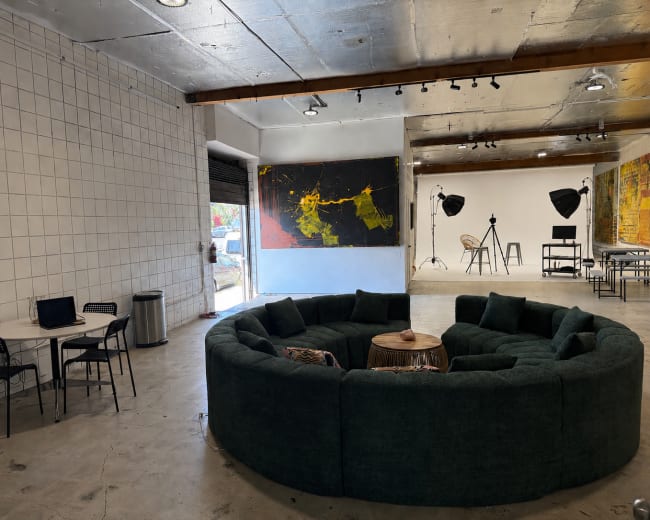 The image shows a spacious interior with a large, circular green sofa, a wooden coffee table in the center, and a dining area with a small table and chairs, while art pieces and photography equipment are visible in the background.