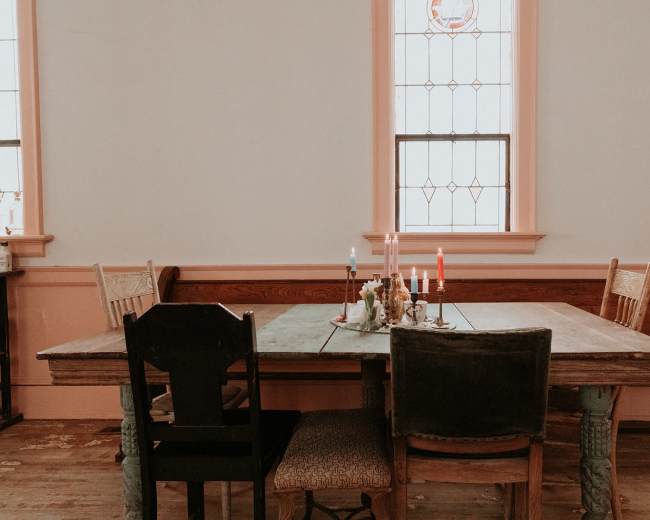 A rustic wooden dining table with various colored candles sits in a room featuring stained glass windows and wooden benches.
