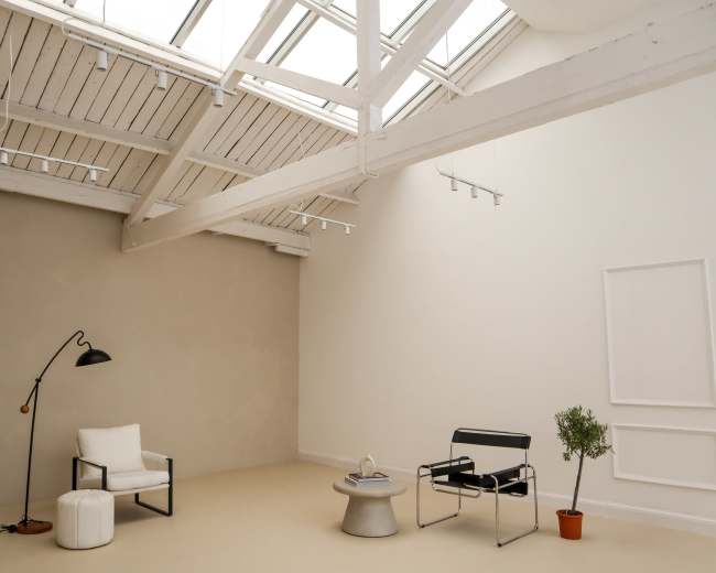 The image shows a minimalist interior space featuring a tall ceiling with skylights, a sofa, a chair, a side table, and a small plant.