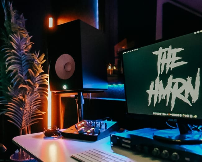 A modern music production studio features a sleek desk with a computer screen displaying "THE AMRN," studio monitors, a keyboard, and DJ equipment, all illuminated by ambient lighting.