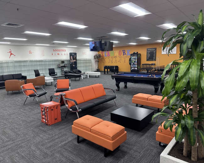 The image shows a modern office lounge area with black and orange seating, a pool table, and a decorative plant.