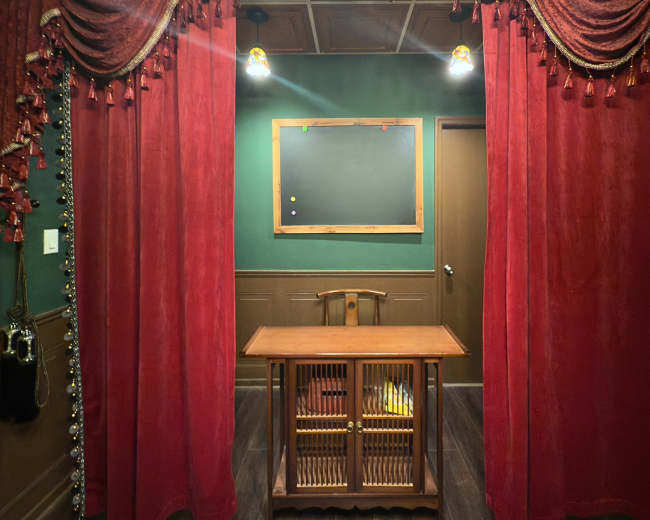 The image shows a room with red curtains framing a wooden table and a chalkboard on a green wall.