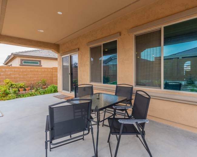 The image shows a patio area with a glass-top table and several black chairs, set against a backdrop of a beige stucco wall and greenery.
