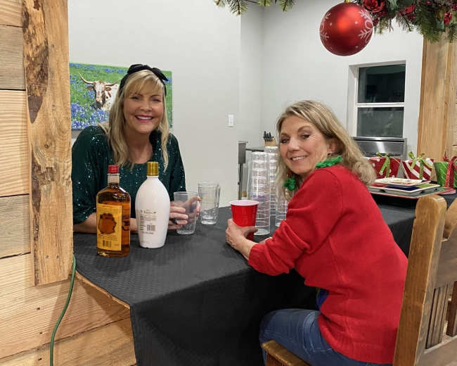Two women sit at a bar counter decorated with holiday ornaments, smiling as they enjoy drinks.
