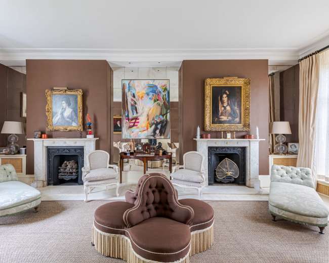 The living room features a symmetrical arrangement of vintage chairs, two ornate paintings on the walls, and a central upholstered sofa on a woven rug.