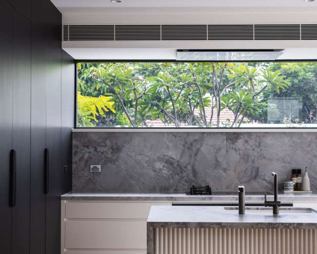 The image shows a modern kitchen with a central island, featuring a marble countertop and a large window overlooking greenery.