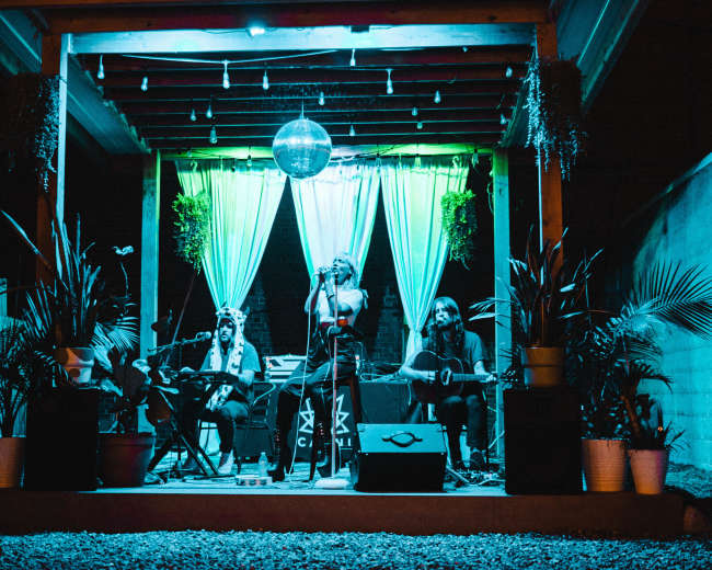 A live music performance takes place on a stage adorned with plants and colorful lighting, featuring musicians playing instruments under a disco ball.