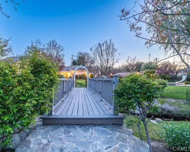 A wooden footbridge leads over a stone path toward a home framed by trees and shrubs, with a soft twilight sky in the background.