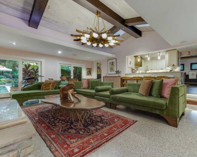 The image shows a modern living room with a green sectional sofa, a wooden coffee table, and a view into an open kitchen area with large windows overlooking a garden.