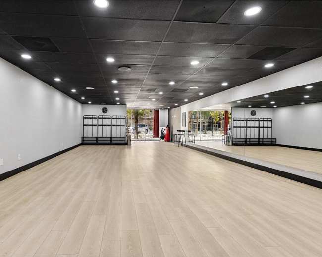 The image shows a spacious, empty room with wooden flooring, mirrored walls, and black ceiling tiles, illuminated by recessed lights.