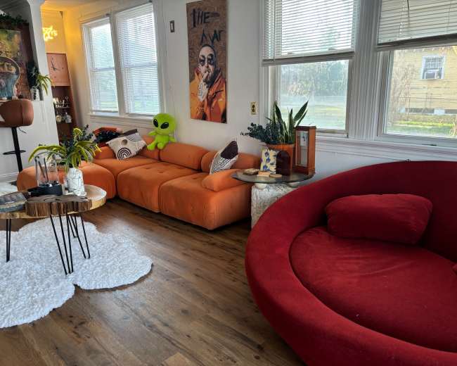 The image features a living room with orange and red sofas, a round coffee table, and a green alien plush toy, with large windows allowing natural light to enter.