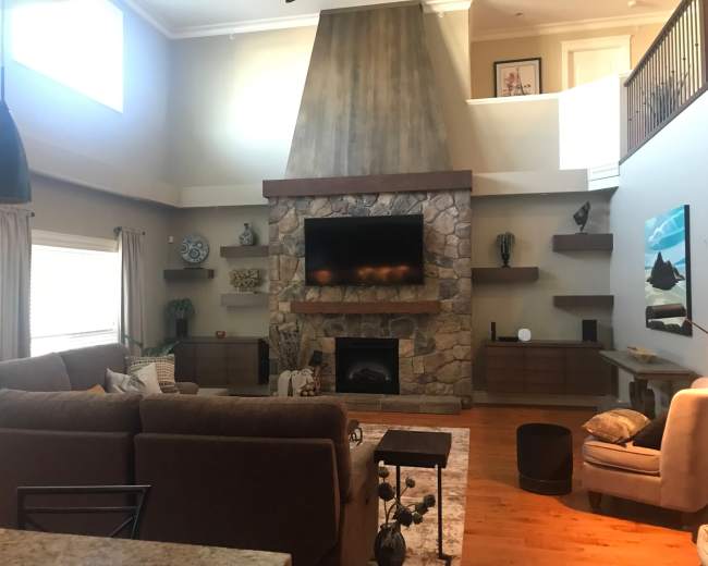 The image shows a spacious living room with a stone fireplace, a large flat-screen TV mounted above it, and a sectional sofa facing the fireplace.