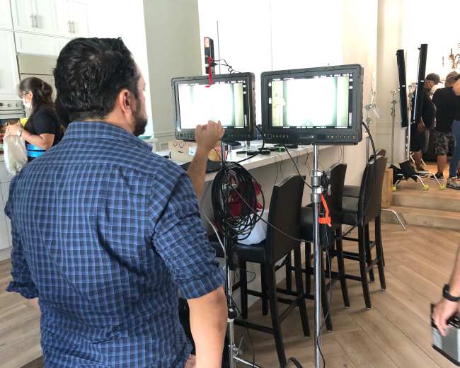 A man adjusts video monitors on a film set while crew members work in the background.