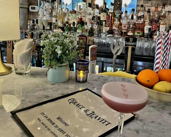 A pink cocktail sits on a marble bar counter with a menu in front of it, while a variety of liquor bottles are visible on shelves in the background.
