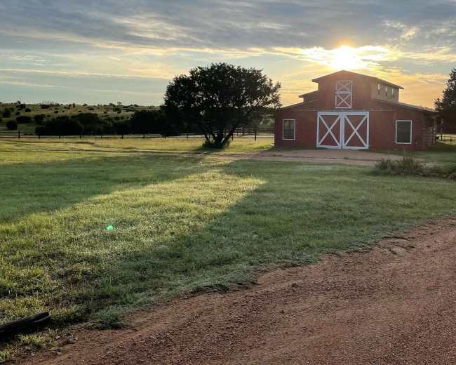 A red barn stands beside a grassy field under a sunrise with scattered clouds in the sky.