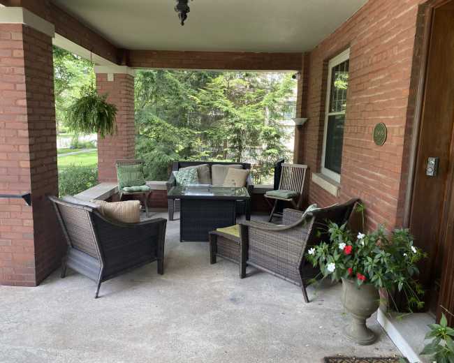 The image shows a porch with a set of wicker furniture, including a coffee table and several chairs, surrounded by greenery.